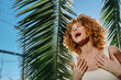 © SHOTPRIME STUDIO - Laughing woman with curly hair enjoys sunny day outdoors near lush palm leaves, expressing happiness and natural beauty under clear blue sky.