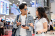 © bongkarn - Asian tourist couple going on trip man holding phone woman talking waiting for departure in airport.
