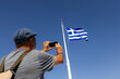 © Jimena Roquero/Stocksy - Tourist taking picture of Greece flag over blue sky on his smartphone