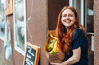 © Westend61 - Happy redhead woman standing with bag of groceries outside store