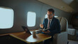 © Videophilia - Businessman in a formal suit reviewing notes in a private jet cabin, focusing on his work with a laptop and coffee at his side while traveling for business