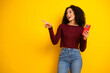 © deagreez - Young mixed race woman with curly hair in a maroon sweater points excitedly while holding a red smartphone against a bright yellow background