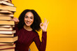 © deagreez - Young mixed race woman with a tall stack of book smiles warmly and makes an ok gesture against a bright yellow background showcasing learning style and positive energy