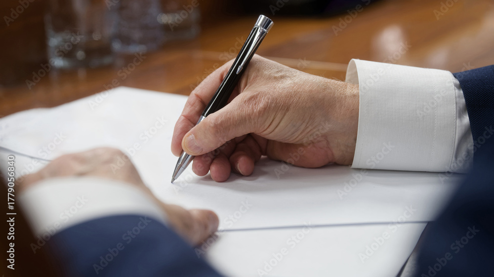 Close-up of an elderly businessman’s hand in a suit writing with a ...