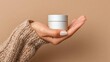 © Anastasiia - A hand in a chunky knit sweater holds a small white cosmetic jar with a lid against a beige background. Concept Hand in chunky knit sweater, Small white cosmetic jar, Beige background
