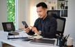 © wattana - Confident businessman using smartphone at modern office desk with laptop and documents.