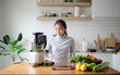 © wattana - Young woman making fresh green juice with a slow juicer in a bright home kitchen.