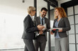 © Novak - Three business professionals standing in a modern office hallway, engaged in discussion while holding a laptop and documents.