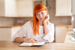 © Iryna - Young woman with red hair contemplates while writing in a notebook in a cozy kitchen setting