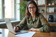 © miss irine - Young Latin woman army soldier works in home office. Smiles at camera, writing documents, using laptop. Military female student wears uniform, admin tasks, online study. Confident professional