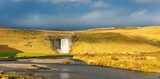 Skogafoss waterfall in Iceland cascades down a steep cliff, creating a powerful mist that forms a vibrant rainbow. The surrounding lush green landscape enhances its majestic beauty