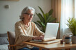 © miss irine - Senior woman uses a laptop computer at home. She types on the keyboard, engaging with online content. Elderly person with gray hair wears glasses, sitting at table. Mature female works or studies.