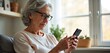 © miss irine - Smiling senior woman uses smartphone at home. Elderly lady texts friends, family on mobile device. Enjoys modern tech to connect, browse social media content. Happy granny stays active, learning new