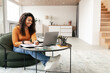 © Prostock-studio - A young woman sits comfortably on a chair while studying. She writes notes and works on her laptop in a cozy, well-lit room with a stylish design.