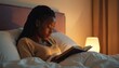 © Viktor - Young African woman reads book in bed before sleeping at night. Black female student relaxes with novel in cozy bedroom under warm lamplight. Girl enjoys literature, studies textbook at home for