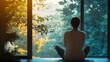 © Crafted - A woman sits in a yoga pose in front of a window, looking out at a forest. The sun shines through the trees, creating a warm glow.