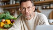 © liliyabatyrova - A remote worker wearing glasses is intently looking at his laptop. Surrounded by fresh vegetables, he sits in a welcoming kitchen filled with natural light, creating a productive atmosphere