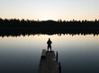 © Dmitri - Aerial drone view of a person's silhouette on a wooden pier overlooking a tranquil Estonian lake with perfect forest reflections at sunset.