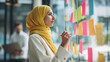 © Katerina Bond - Confident businesswoman in yellow hijab writing on colorful sticky notes during brainstorming session in modern office, planning and organizing ideas for team project.