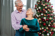 © Юлия Завалишина - Merry Christmas. Happy senior couple smiling near Christmas tree at home. Happy family old man woman with Christmas tree. Grandparents celebrating New Year together near tree fir. Family moments