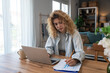 © Srdjan - Young woman working from home on laptop casual striped shirt and coffee nearby, reviewing charts and typing with calm focus remote work productivity and budgeting at wooden table in cozy living room
