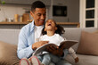 © Home-stock - Storytime. Happy loving black father reading fairy tales to his cute little daughter, sitting together with big book on sofa and girl laughing
