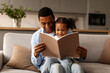 © Home-stock - Loving African American father and his little daughter reading book together or looking photo album, girl sitting on daddy's laps on sofa in living room