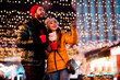 © deagreez - Joyful couple under festive lights enjoys warm drinks at night market celebrating christmas magic together