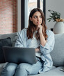 © Daniel - Distracted from work worried young woman sitting on couch with laptop, thinking of problems. Pensive unmotivated lady looking at window, feeling lack of energy, doing remote freelance tasks at home.