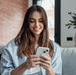 © Daniel - Smiling woman looking at smartphone screen, sitting on couch, enjoying leisure time with mobile device at home, happy young female chatting in social network or surfing internet, shopping online