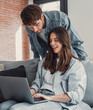 © Daniel - Happy millennial couple sit relax on couch in living room, smiling young husband standing from back and wife rest on sofa at home browsing Internet using modern computer device