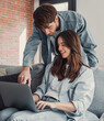 © Daniel - Happy millennial couple sit relax on couch in living room, smiling young husband standing from back and wife rest on sofa at home browsing Internet using modern computer device