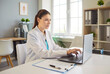 © Studio Romantic - Young female doctor in white coat sits at desk and working on laptop in modern clinic office, checking patient data or medical records. Online consultations, digital health records management concept