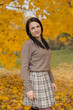 © Татьяна Зернова - A young Caucasian woman with long dark hair stands in a park surrounded by vibrant autumn leaves. She wears a brown sweater and a plaid skirt.
