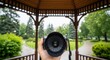 © Mariya Sorvacheva - First-person view of a hand holding a camera with a rain-speckled lens, capturing lush greenery in a serene park gazebo setting, showcasing nature photography and artistic expression
