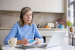 © Liubomir - Senior woman engaging in remote work or e-learning from her home kitchen, wearing a headset, taking notes in a notebook while looking at her laptop screen