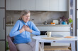 © Liubomir - Senior woman sitting at a kitchen table, grabbing her chest with both hands, expressing a look of intense pain and discomfort, suggesting a heart attack or acute health issue