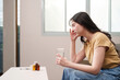 © Natee Meepian - Health and Wellness. Young woman with headache at home, holding glass of water.