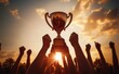 © Tatyana - Multiple hands raised in celebration holding a large trophy against a golden sunset sky expressing victory and achievement