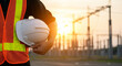 © mod - A construction worker holding a white hard hat in front of an electrical substation at sunset time