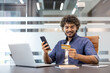 © Tetiana - Smiling young Indian man sitting at office desk, using mobile phone and credit card