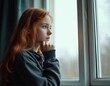 © miss irine - Pensive redhead girl looks through window at home. Teenager deep in thought with sad eyes. Young female contemplates her future during solitude in quarantine. Kid feels depressed indoors
