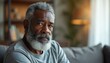 © miss irine - Mature African American man with grey beard sits on sofa. He looks directly ahead with a serious thoughtful expression. His face shows wrinkles and experience. He wears simple gray shirt.