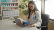 © Krakenimages.com - Young chinese woman doctor in a clinic room, wearing a stethoscope and uniform, works on a laptop and tablet, surrounded by medical supplies and documents.