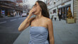 © Krakenimages.com - Woman covering mouth while yawning on a city street, strapless dress revealing shoulder and collarbone, pedestrians blurred nearby; sleepy.