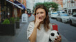 © Krakenimages.com - Woman holding soccer ball and gesturing peace sign on busy city street with cars passing by in urban outdoor setting during daytime, wearing casual white shirt.