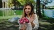 © Krakenimages.com - Woman holding flowers in a park with vibrant green trees, wearing a white shirt with a tattoo on her arm, expressing gratitude and happiness under sunny weather.