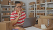 © Krakenimages.com - Man typing on laptop and points finger to laptop inside building filled with stacked parcels and shelves while checking labels and tape; focus productivity.