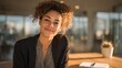 © Luan - Smiling woman with curly hair and blazer sitting at a table in a bright modern space inside