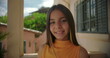 © Marco - Close-up portrait of a smiling young girl on a Latin American balcony with pastel buildings and greenery in the background, cheerful and confident expression
